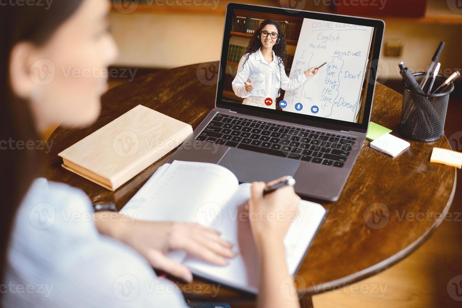 E-learning Concept. Over the shoulder back view of female student sitting at table, writing in notebook, taking notes, using laptop, having online class with English teacher. Distant studying photo