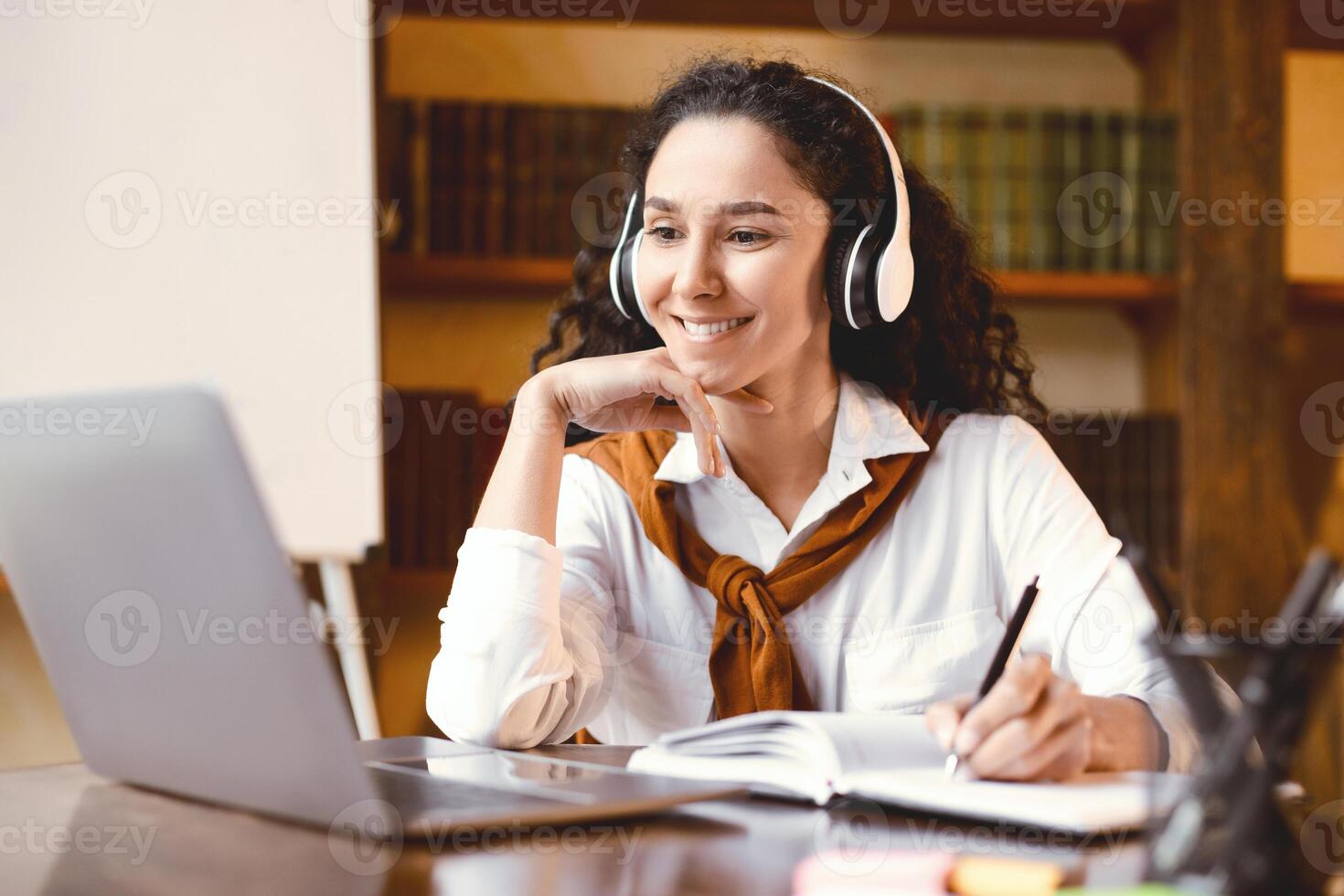 Doing Homework. Portrait of smiling young woman sitting at desk with pen, writing and taking notes in notebook, using and looking at laptop computer. Cheerful curly lady watching online course photo