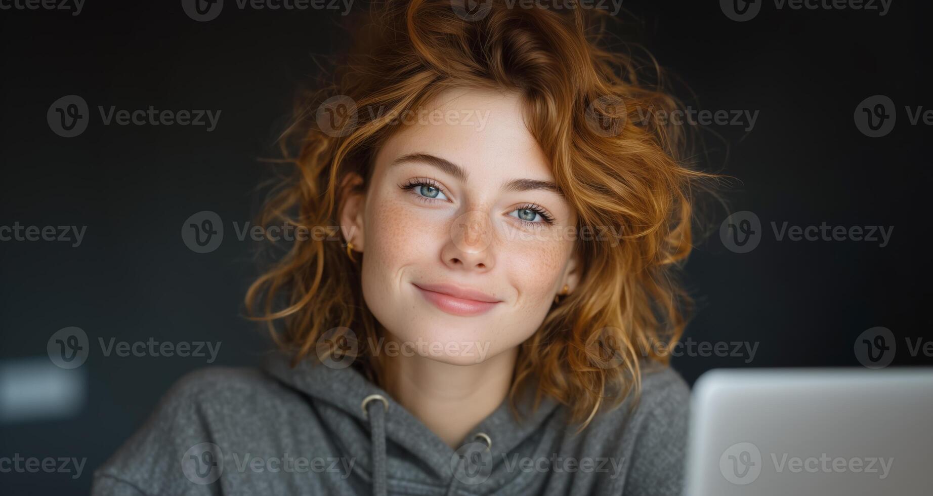 A young woman sits in front of a laptop, smiling while interacting with voice control technology. Her curly hair frames her face as she enjoys a casual moment during her tech-focused activity. photo