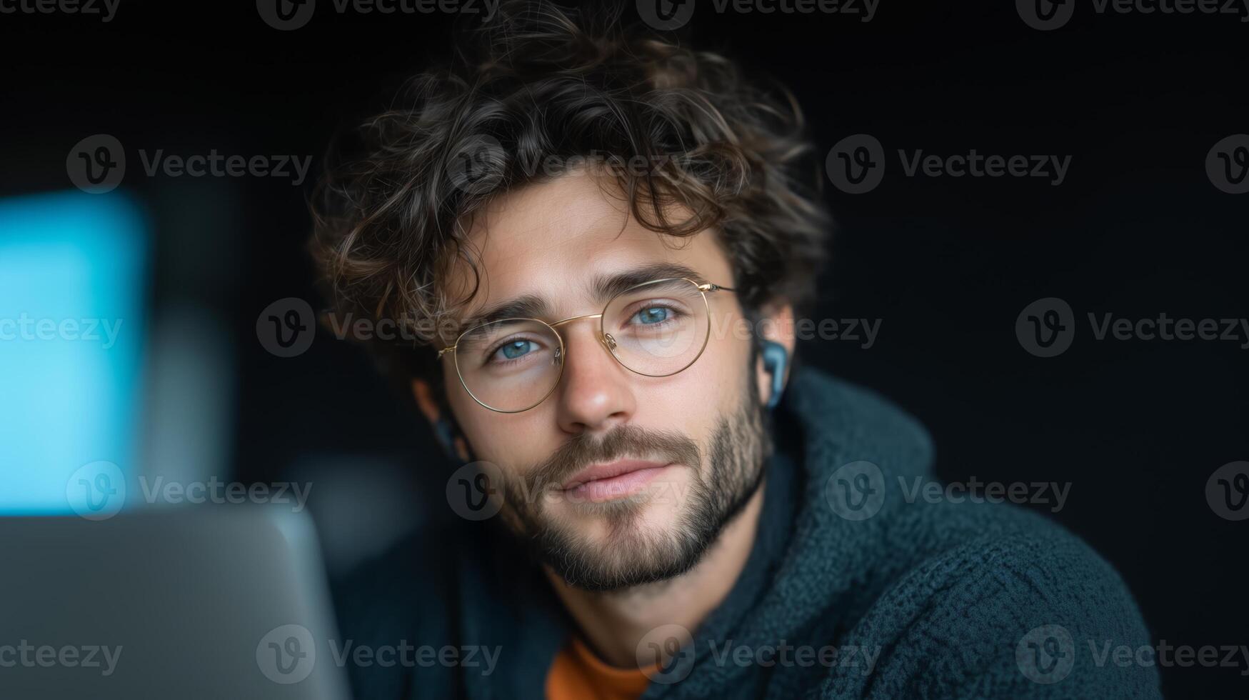 A young man with curly hair and glasses engages with a voice assistant using earbuds while working on a laptop. The setting is cozy, with a soft focus background that emphasizes his concentration. photo