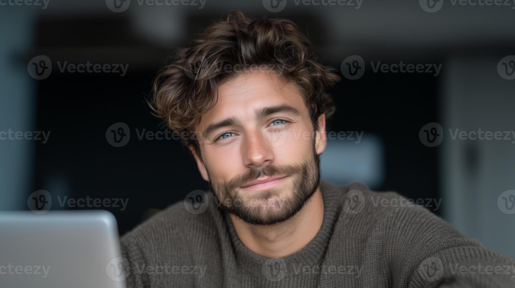 A man with curly hair and a beard smiles while using a laptop in a contemporary workspace. He interacts with a voice assistant, showcasing a seamless connection for communication and productivity. photo