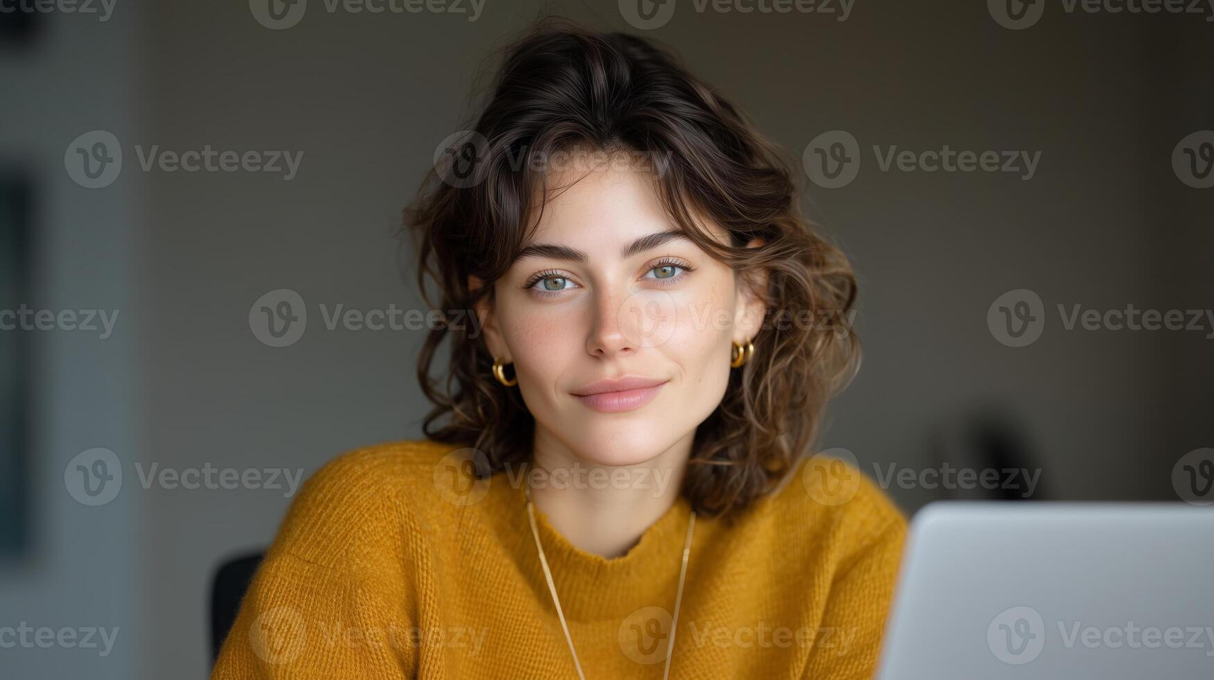 A young woman with curly hair sits at a desk, focused on her laptop. She is interacting with voice control technology, illustrating effective communication while working. photo