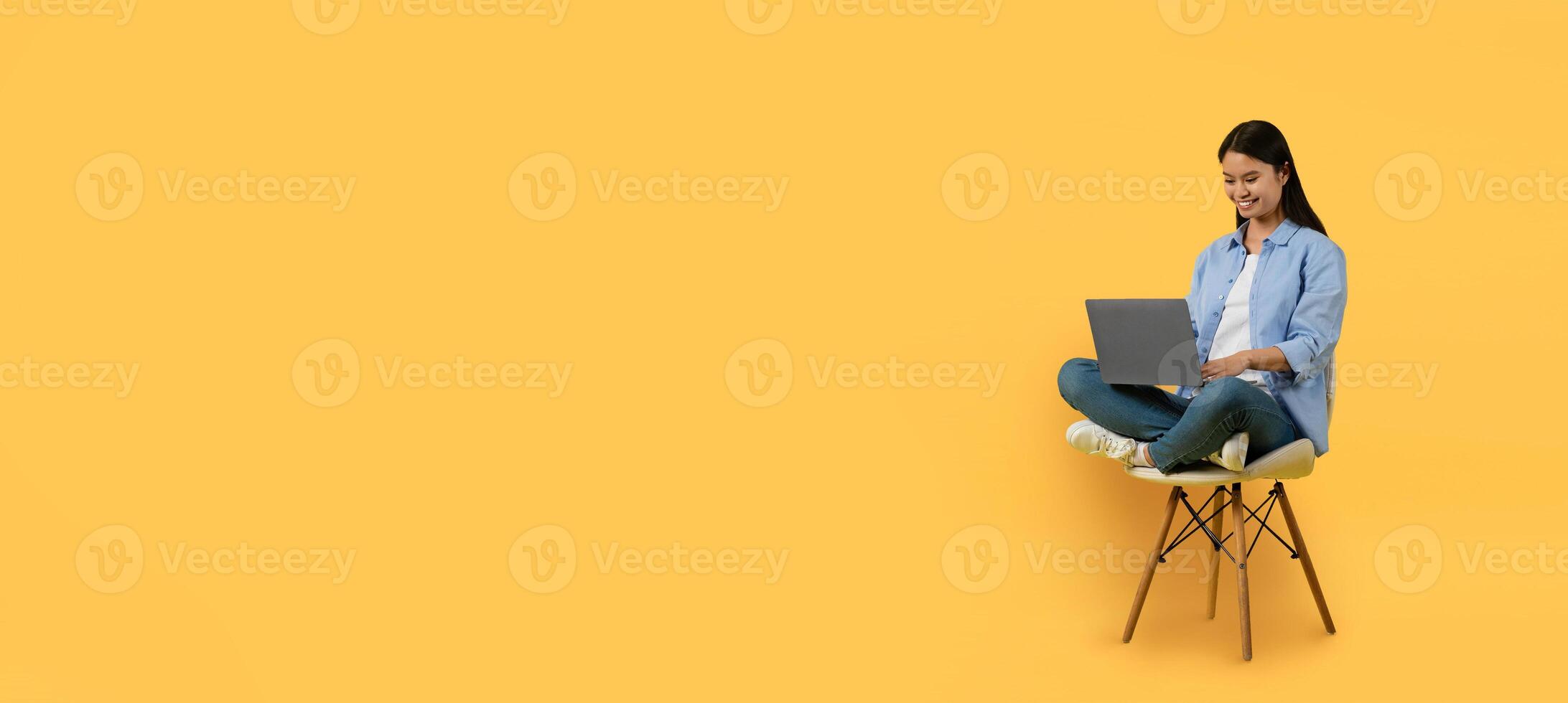Asian young woman smiles while typing on her laptop, sitting cross-legged on a chair. The bright yellow background enhances the cheerful atmosphere of her work environment. photo