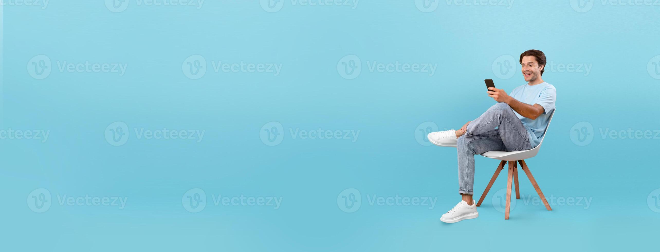 A young man sits casually in a white chair, smiling while using his smartphone. The vibrant blue background enhances the cheerful atmosphere, highlighting his relaxed demeanor. photo