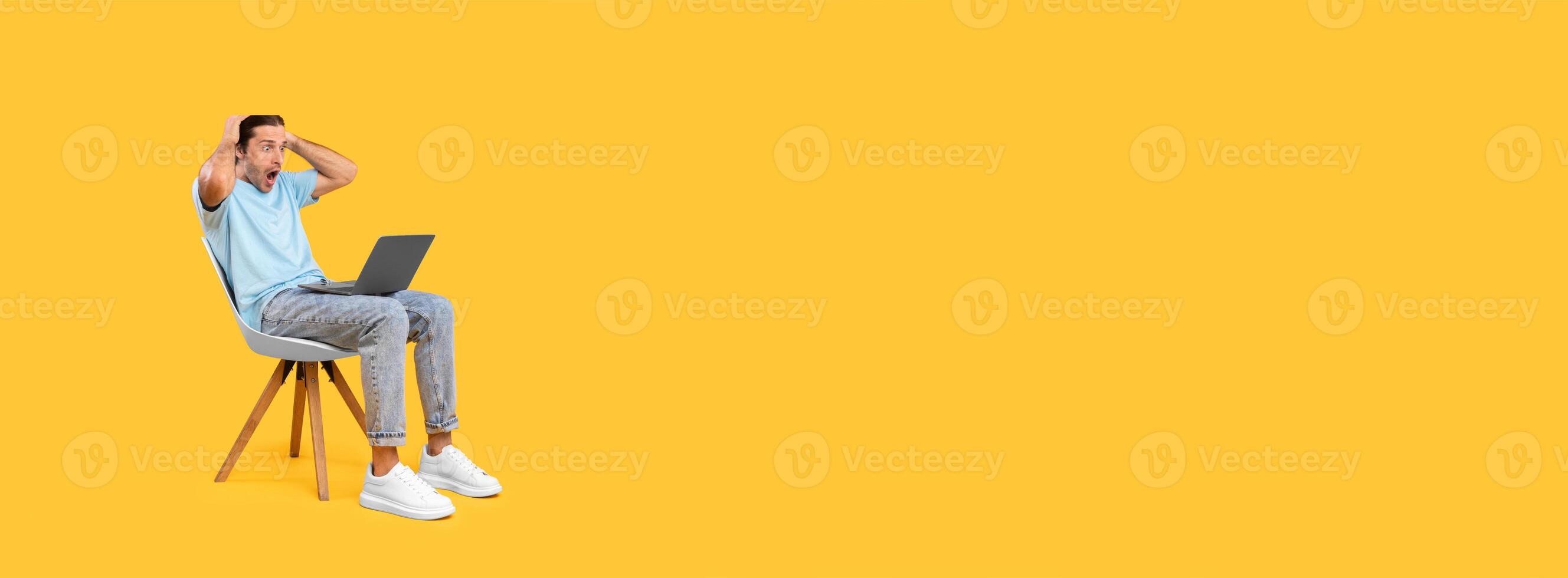 A young man sits on a wooden chair with his hands on his head, appearing stressed while working on a laptop. The vibrant yellow wall creates a bold contrast with his casual outfit. photo