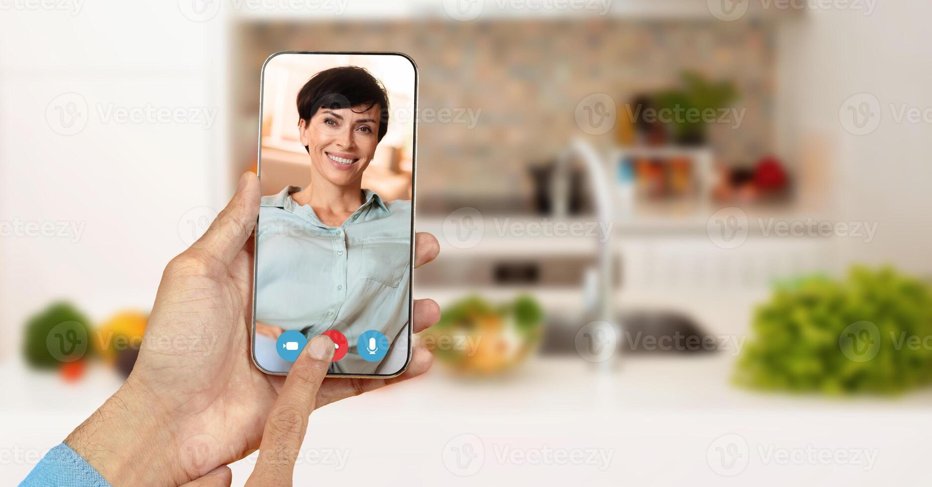 A person holds a smartphone with smiling face during a call in a cozy kitchen. Fresh ingredients and a homey atmosphere create a casual setting for communication and connection. photo