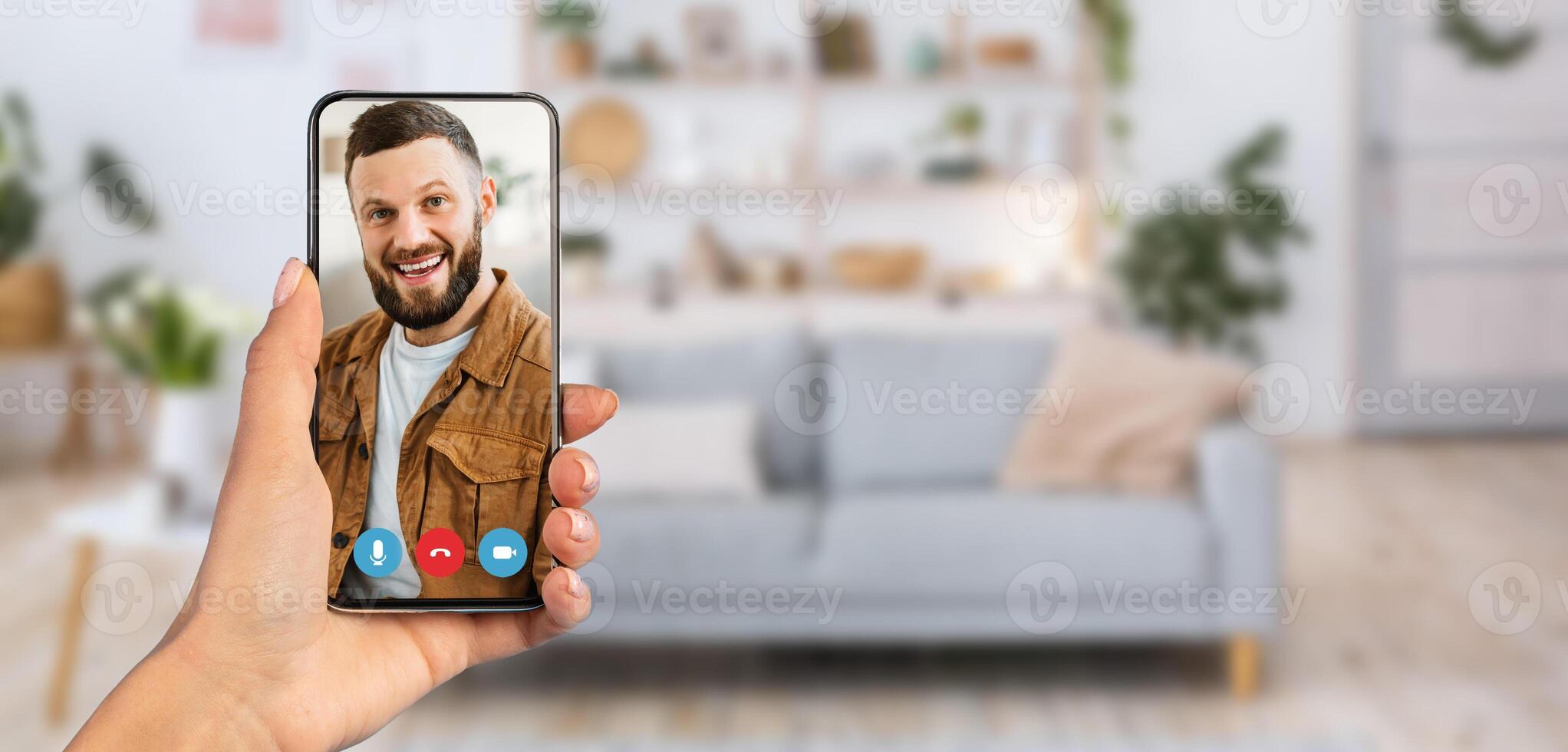 A person is holding a smartphone during a call. The background features a comfortable living room with a sofa, plants, and decorative shelves, creating a relaxed atmosphere. photo
