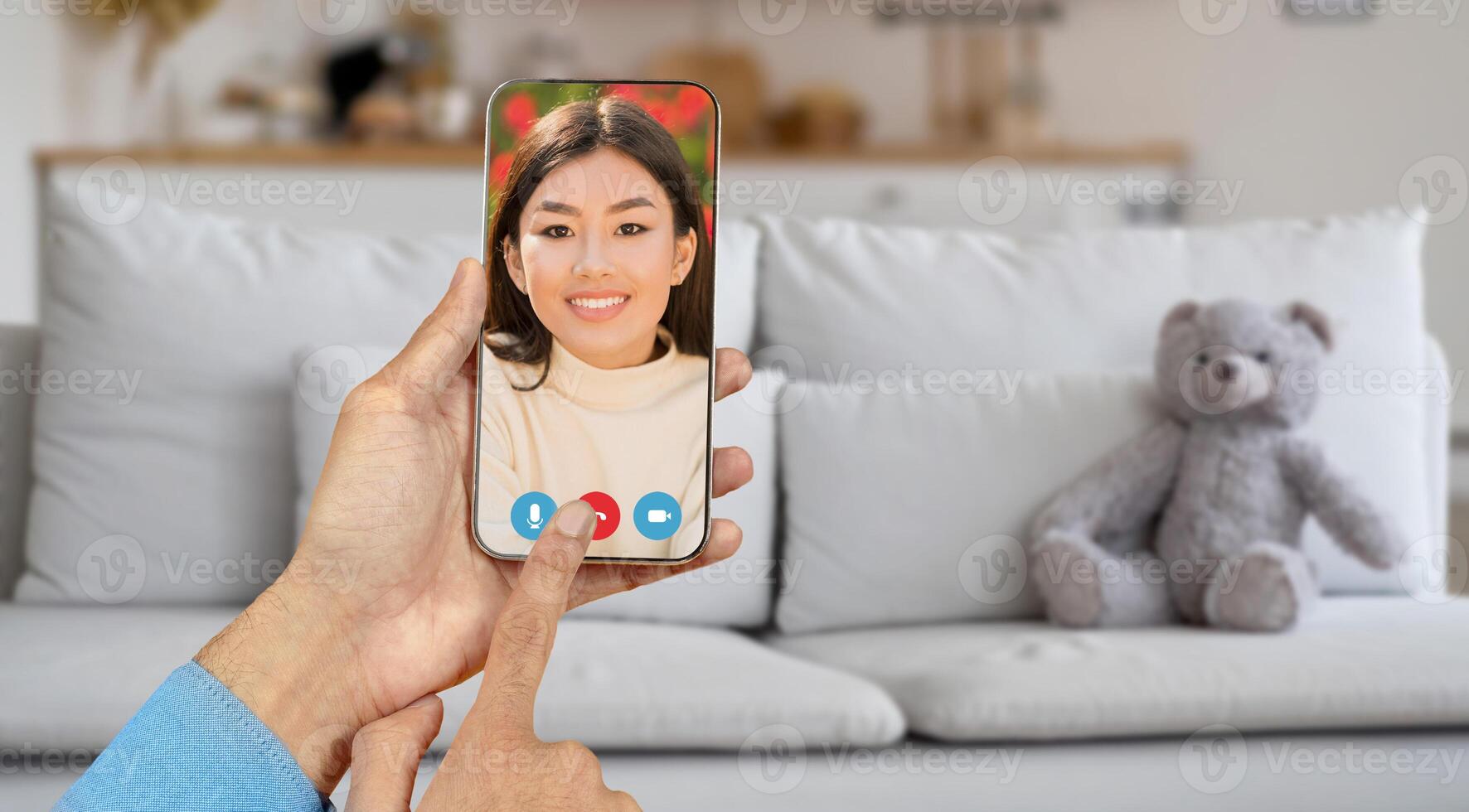 A person holds a smartphone showing a smiling individual on the screen, engaged in a call from a cozy living room with a teddy bear on the couch. photo