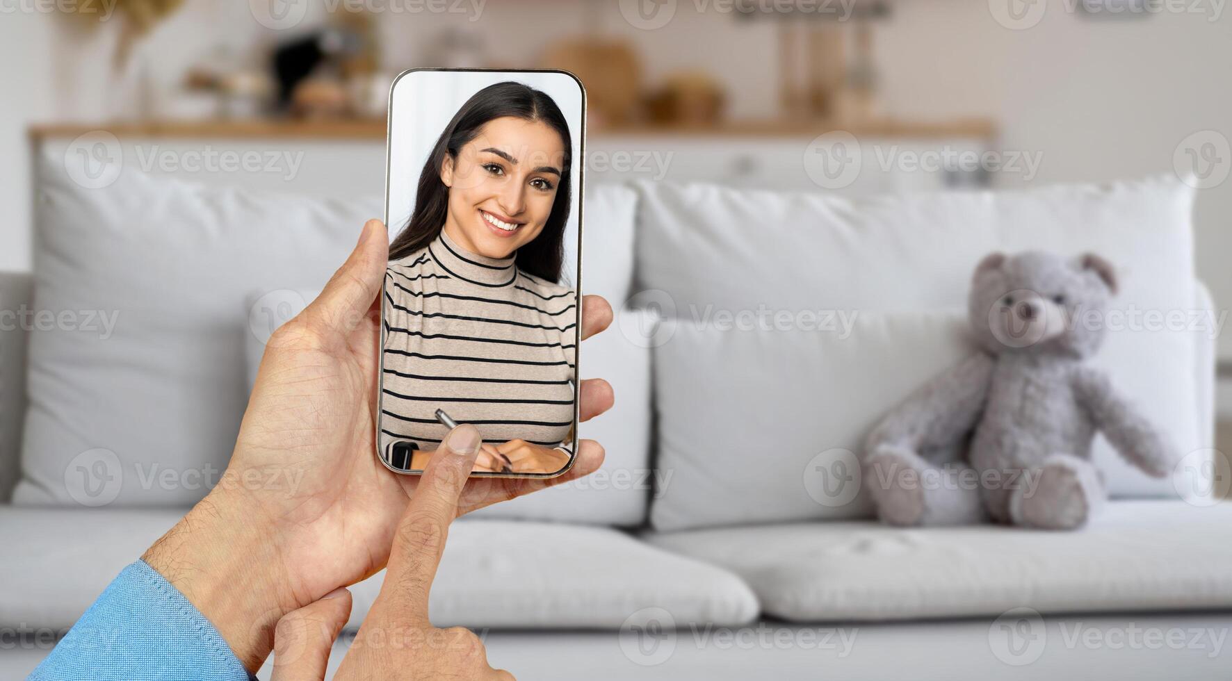 A person interacts with a smiling individual on their smartphone screen while sitting in a cozy living room. The warm atmosphere contributes to an inviting virtual communication experience. photo