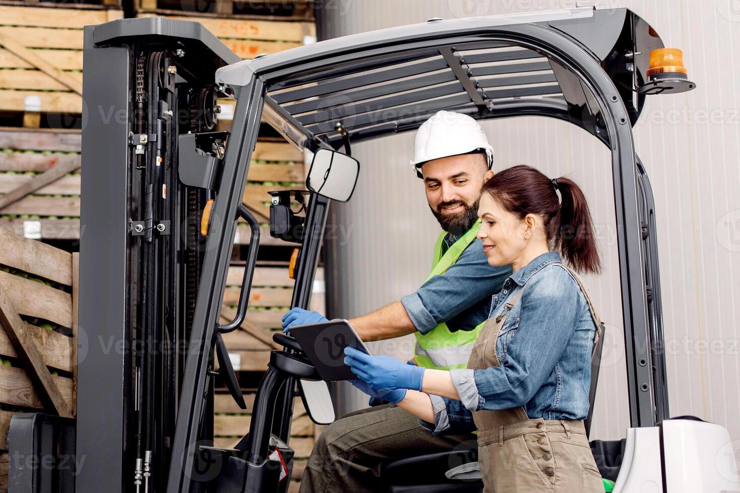 Management of loading products, modern device and checking driver work in warehouse. Cheerful male in helmet lifts up apples in container on shelves with wooden crates, lady control man on fork lift photo