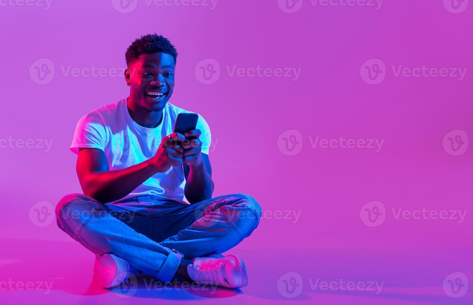 Full length of millennial African American guy sitting cross legged with smartphone, surfing social media, chatting to friend in neon light, copy space. Cool black man using mobile phone photo
