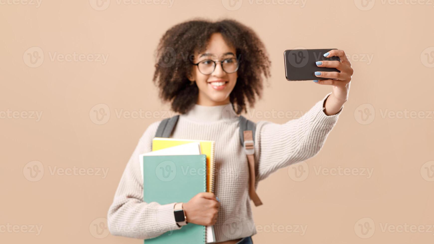 Photo for educational blog. Smiling smart african american young lady in glasses with backpack and smart watch holding notebooks and makes selfie isolated on yellow background, studio shot, copy space