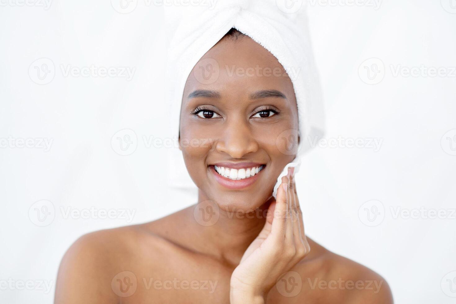 Beauty care, remove makeup and use tonic at home. Attractive fresh young african american lady with white towel on head rubs face with cotton pad and tonic after shower on white background, copy space photo