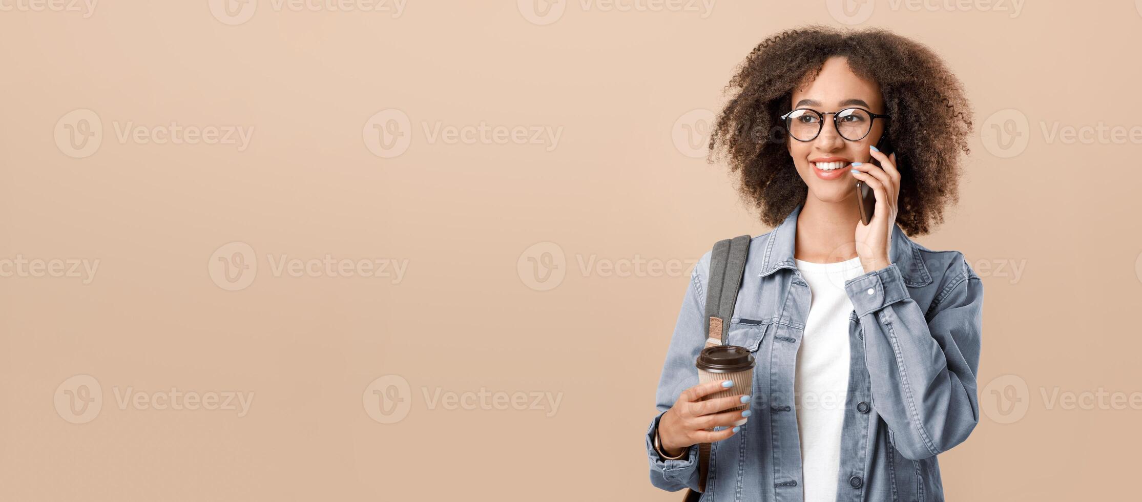 Modern worker, freelancer or blogger. Smiling teen african american curly lady in glasses with backpack, speaks on phone and holds cup of coffee takeaway, isolated on light background, free space photo