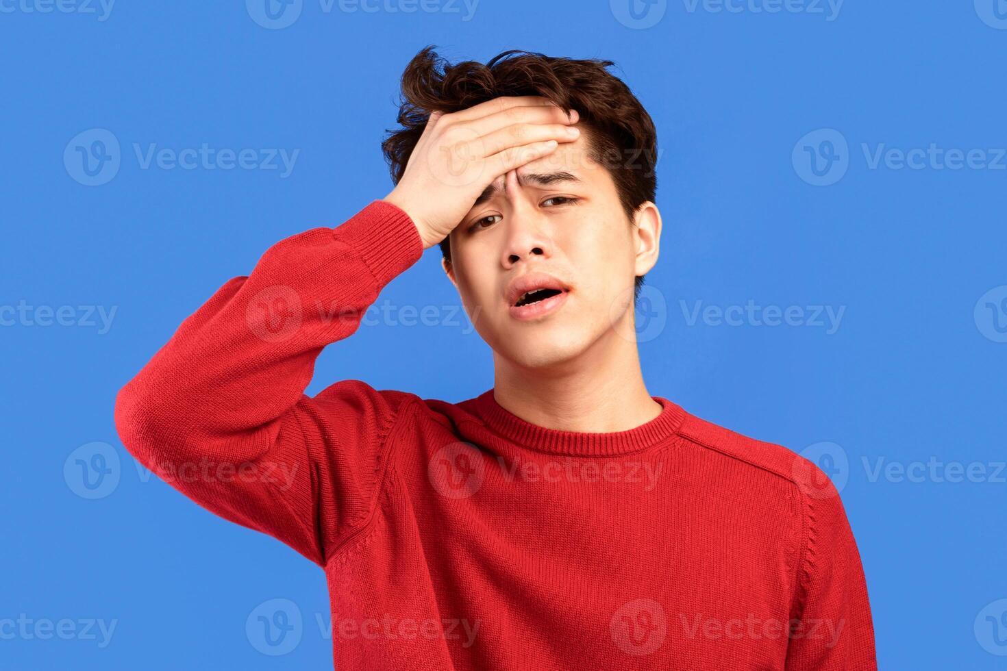 Fever And Headache. Portrait of weak unwell asian man with black hair feeling bad, having high temperature or physical pressure, touching forehead with palm, isolated over blue studio background photo