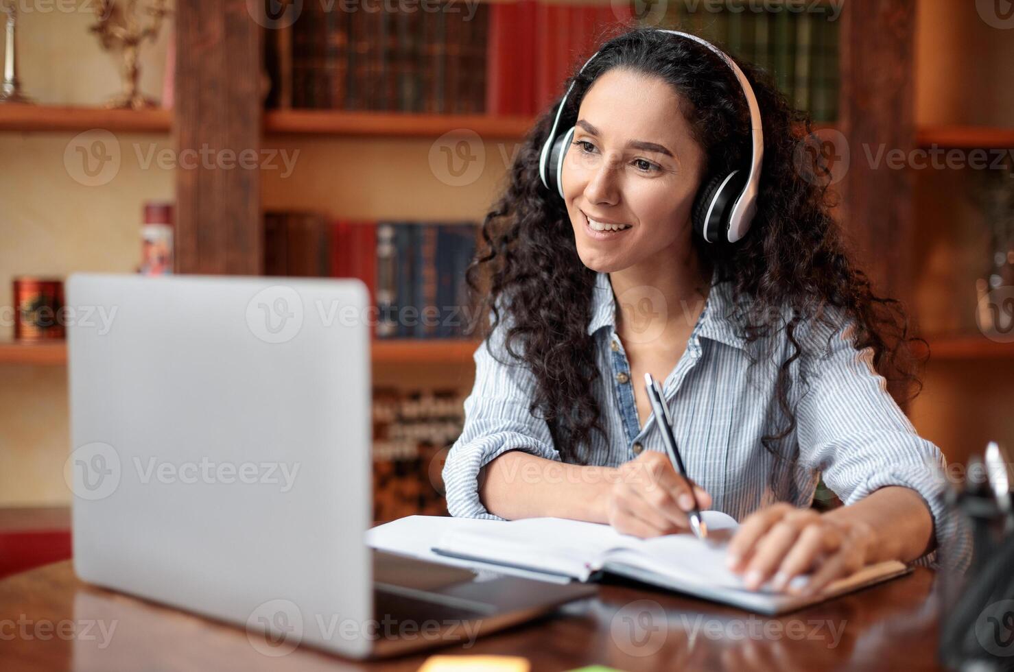 Modern Technology And Elearning. Smiling woman in wireless headset watching webinar online, sitting at desk, using laptop and writing in notebook, taking notes. Education During Quarantine photo