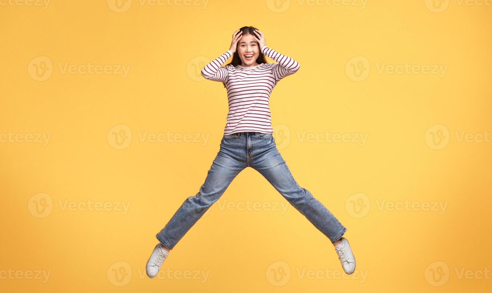 A cheerful young woman leaps joyfully, displaying excitement and energy. She wears a striped sweater and jeans, creating a vibrant contrast against the sunny yellow backdrop. photo