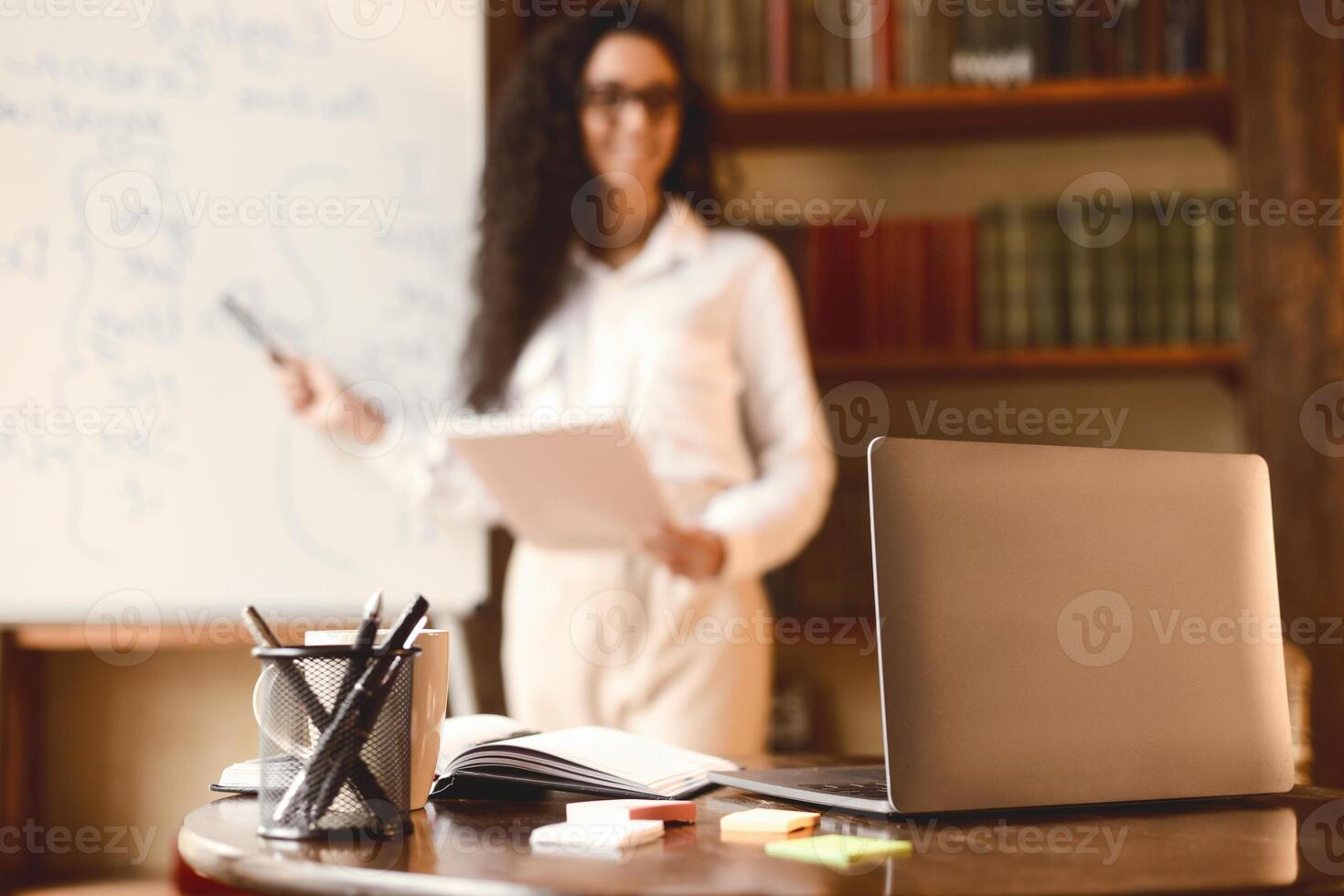 Workspace. Selective focus on laptop on the table, smiling woman in eyeglasses standing at whiteboard in the blurred background. E-learning, remote work, manager giving presentation, online briefing photo