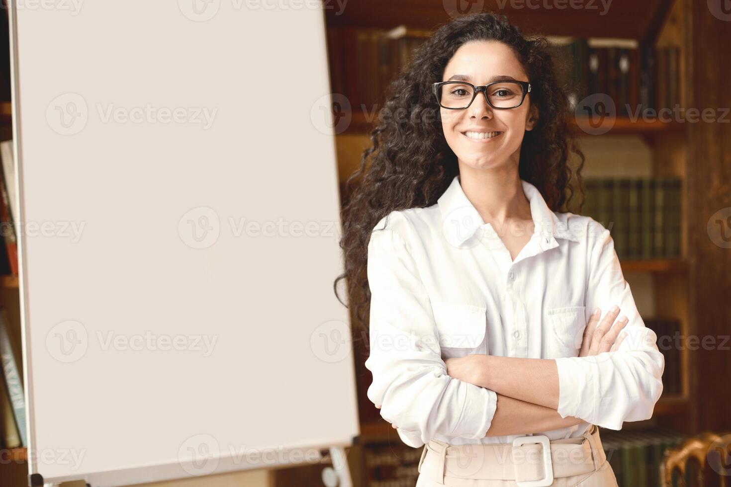Meeting And Education. Portrait of smiling young lady in eyewear standing with folded arms and posing at camera near blank empty white board in the background. Happy casual woman in shirt photo