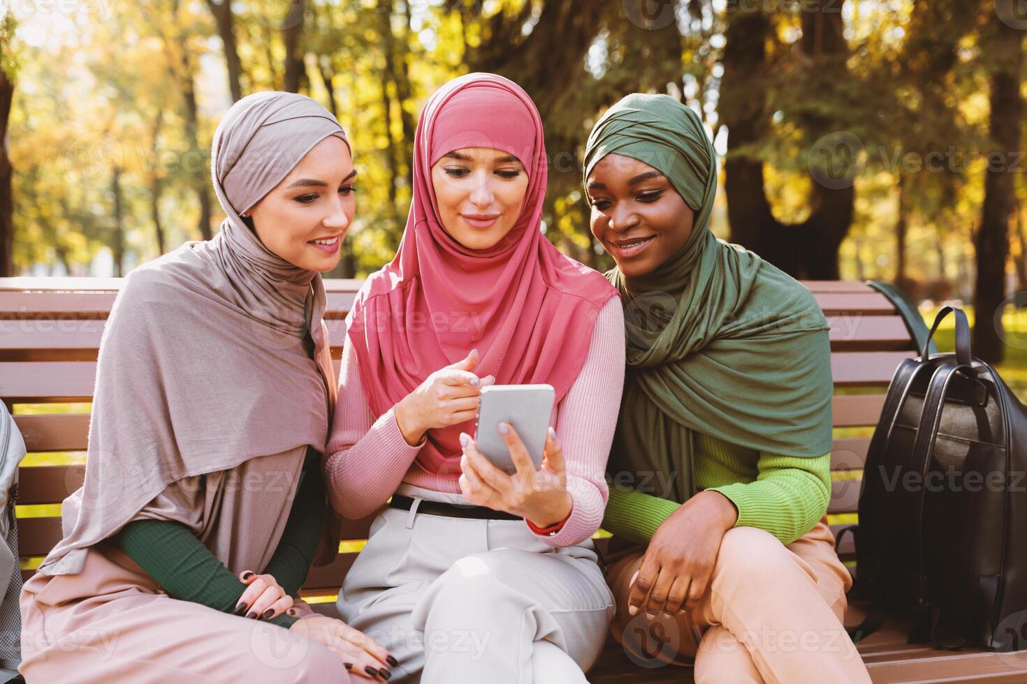 Modern Muslim Students Ladies Using Smartphone With Educational Mobile App Learning Together Sitting On Bench In Park Outdoor. Great Application For Your Cellphone Concept photo