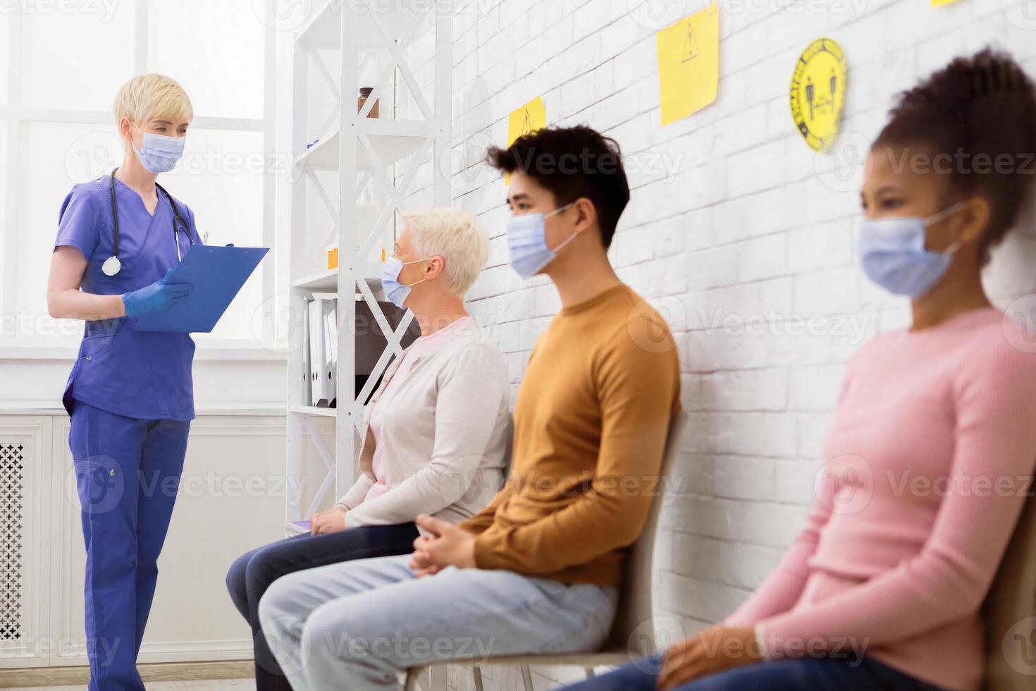 Coronavirus Treatment. Diverse People Sitting In Queue Waiting For Doctor's Appointment, Nurse Talking With Patients In Hospital Waiting-Room. Covid-19 Medical Help And Vaccination. Selective Focus photo