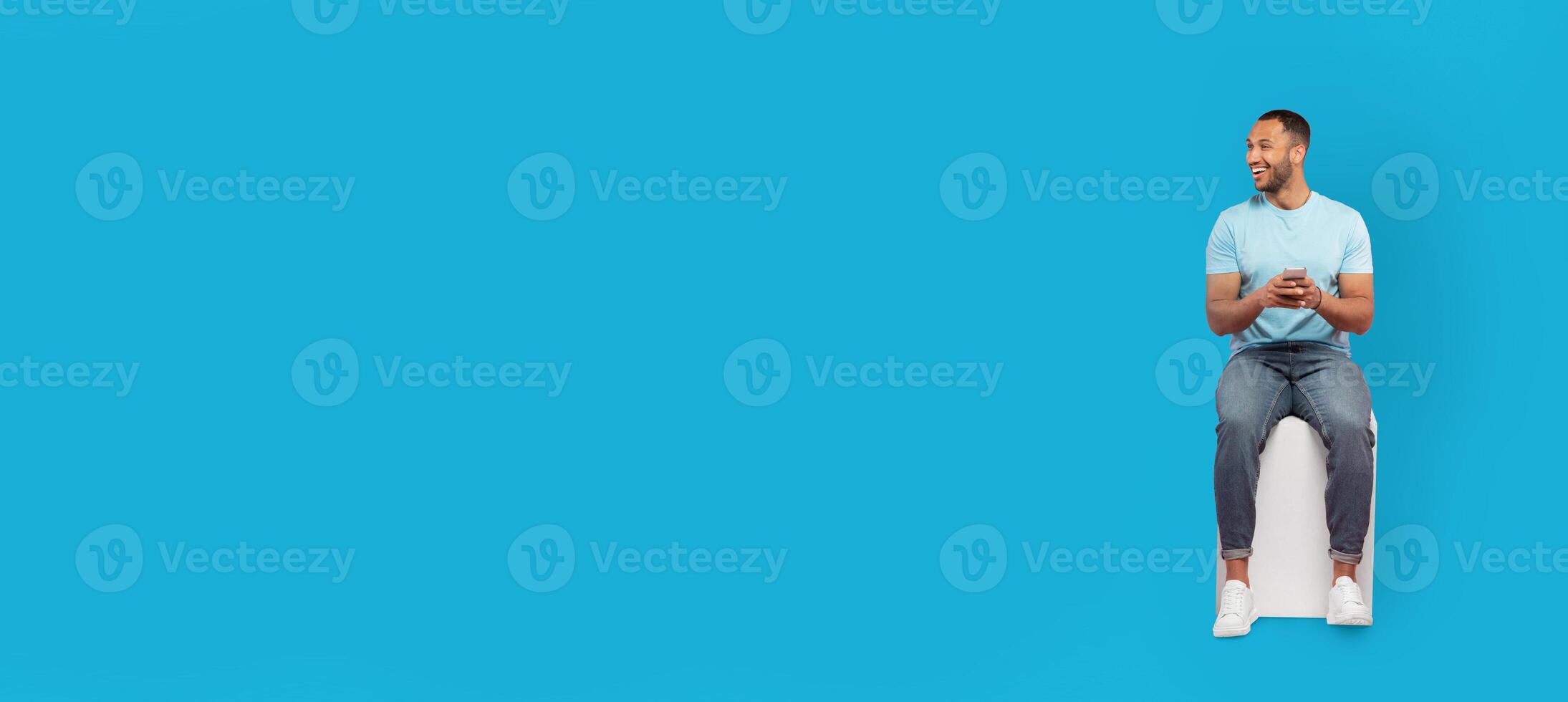 A young African American man in a light blue shirt sits on a white stool, smiling and looking at his phone. The vibrant blue background enhances the cheerful atmosphere. photo