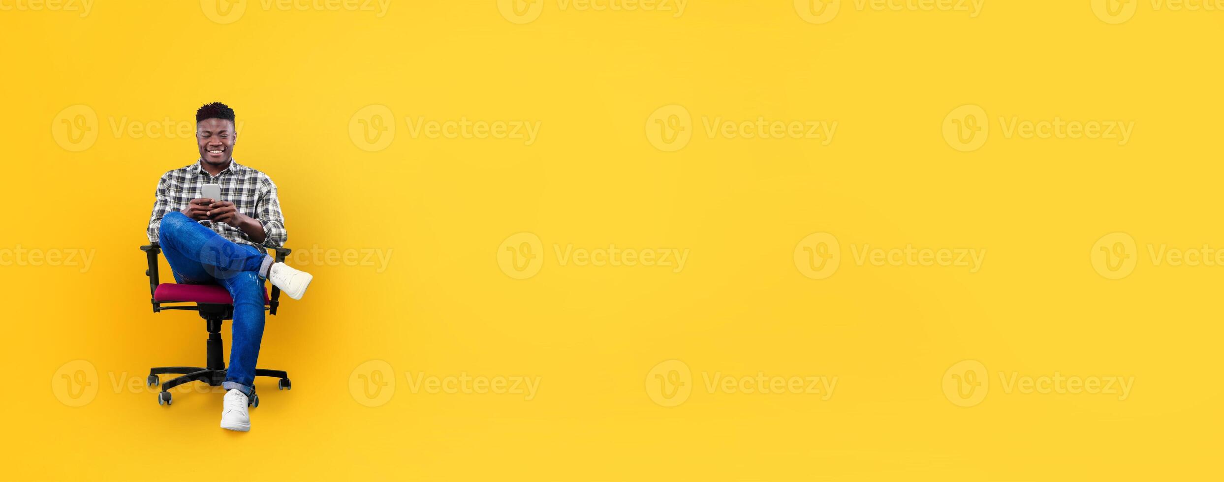African American young man sits comfortably in a black and red chair, casually browsing his smartphone against a vibrant yellow backdrop. He appears relaxed and content in this bright setting. photo