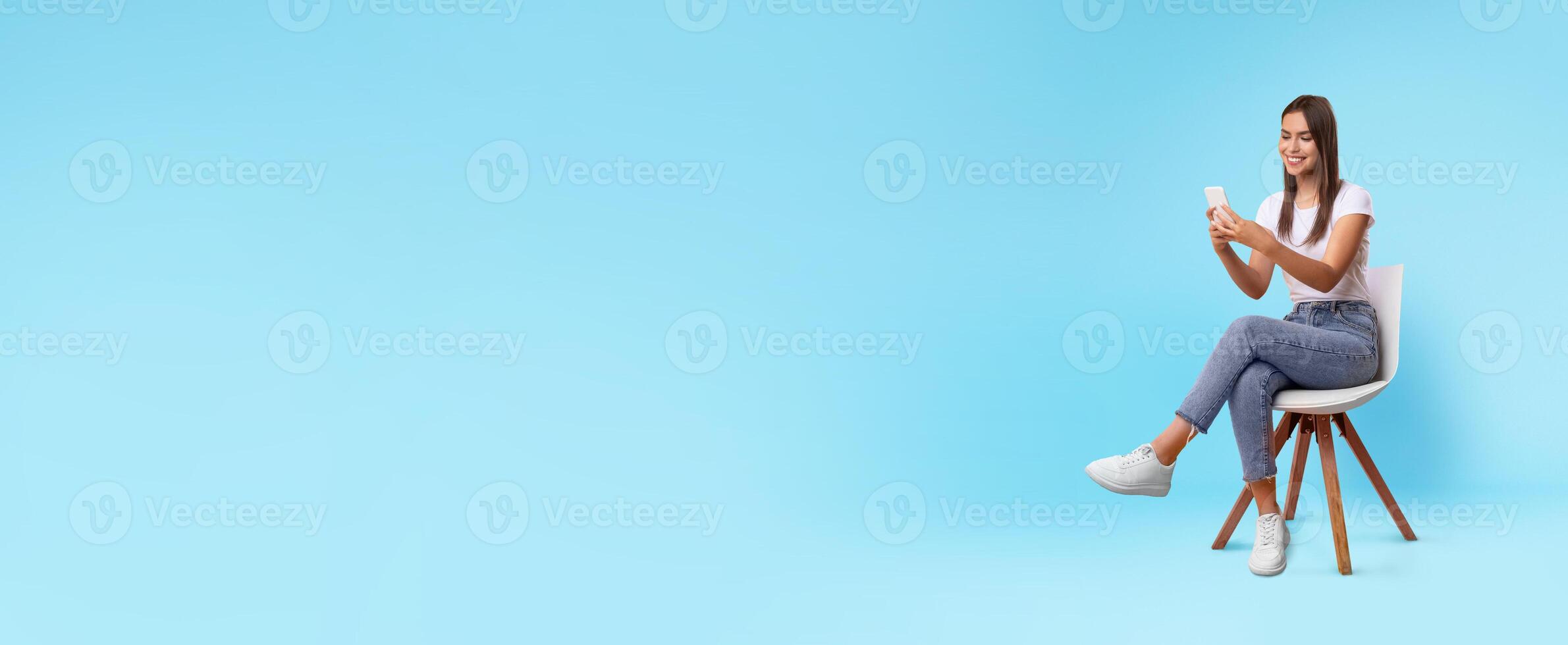 A woman is seated on a chair, focused on her smartphone as she enjoys browsing. The vibrant blue background enhances the modern and casual atmosphere, creating a lively mood. photo