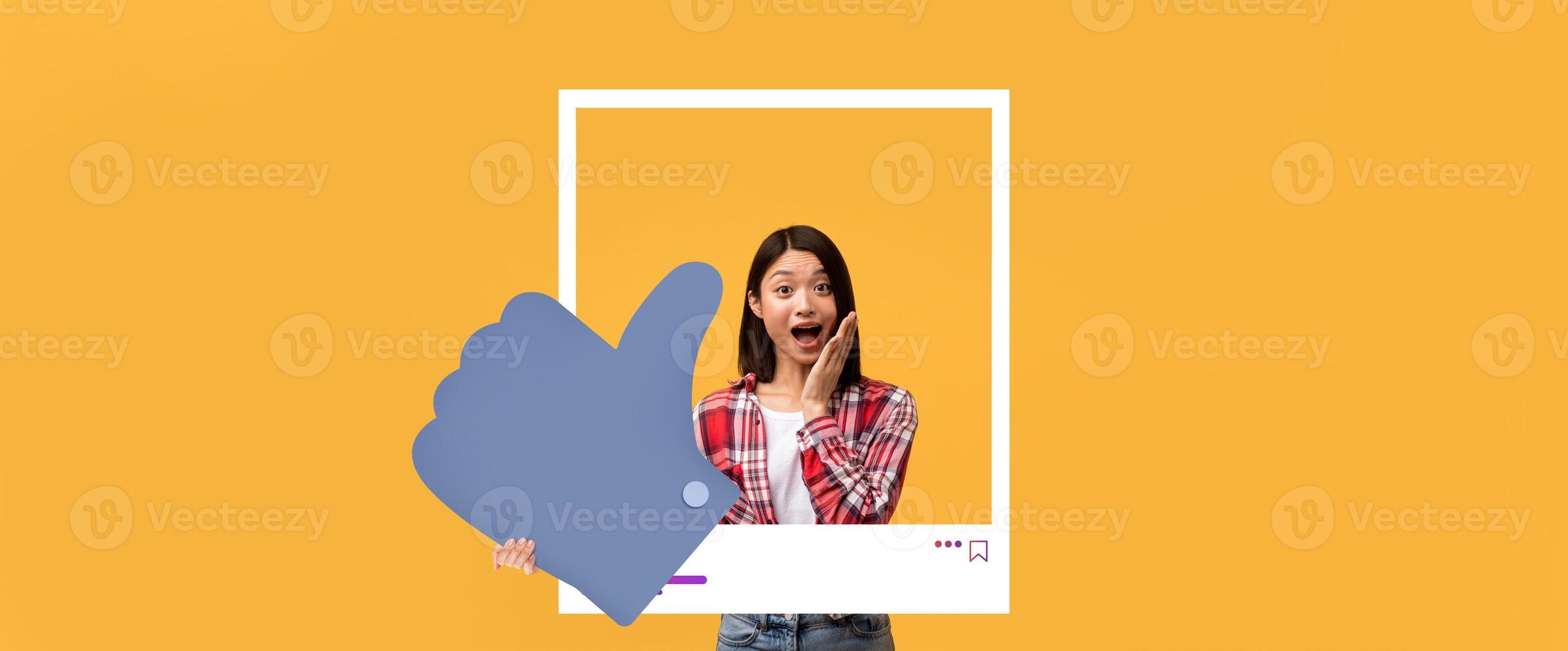 A young woman stands with a surprised expression, holding a large thumbs up near her face. The bright yellow backdrop enhances the playful atmosphere of social media engagement and content creation. photo