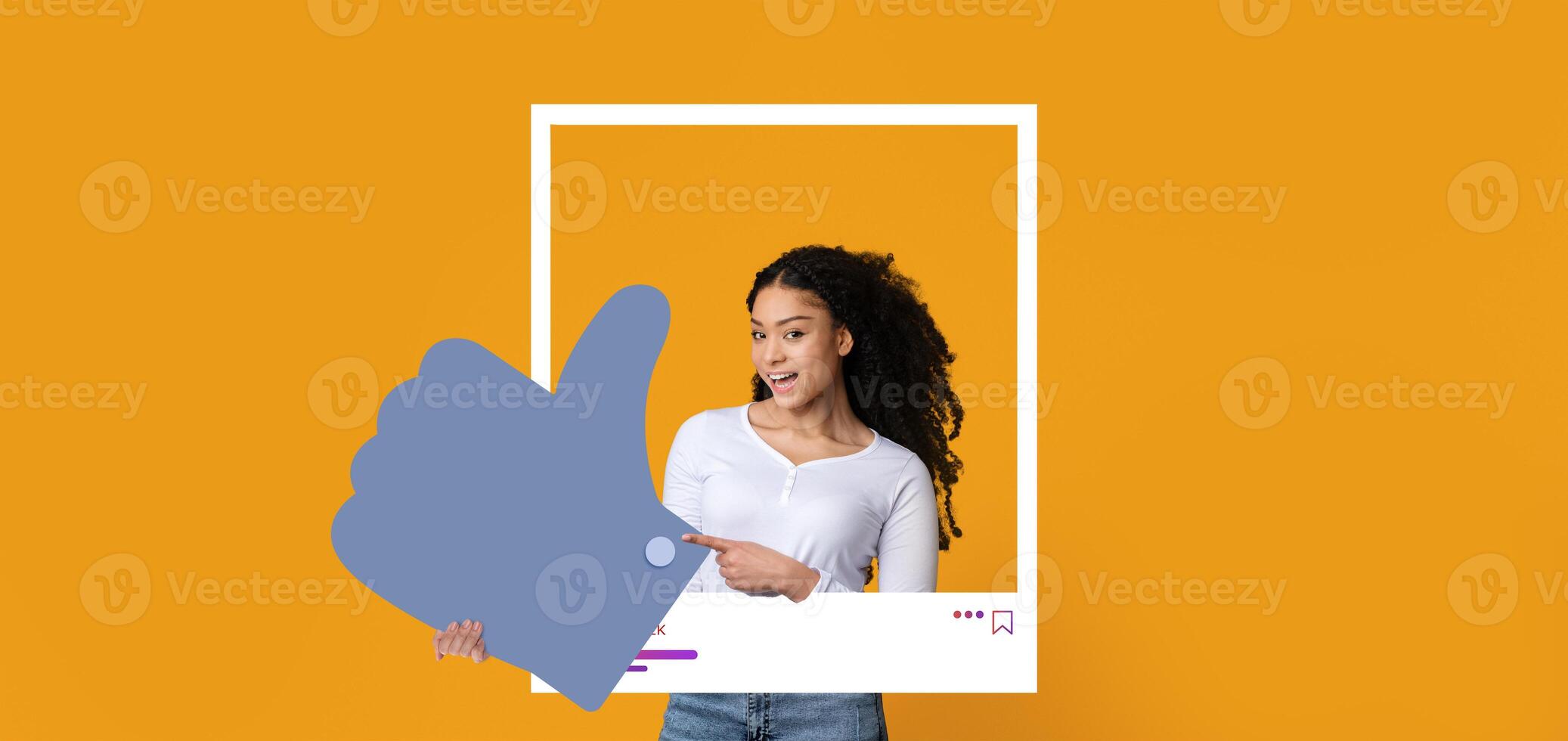 A young woman with curly hair smiles while holding a large thumbs up sign, embodying positivity and engagement in social media interactions against a bright orange backdrop. photo