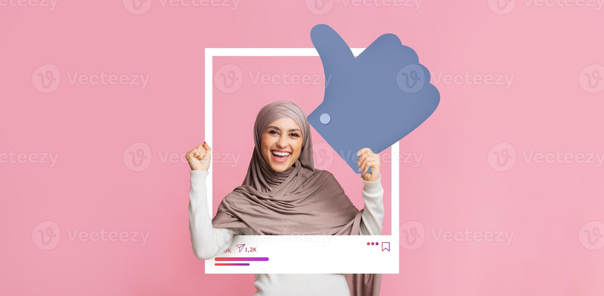 A woman wearing a hijab joyfully poses inside a social media frame, giving a thumbs up to celebrate her content engagement. The bright pink background adds to the lively atmosphere. photo