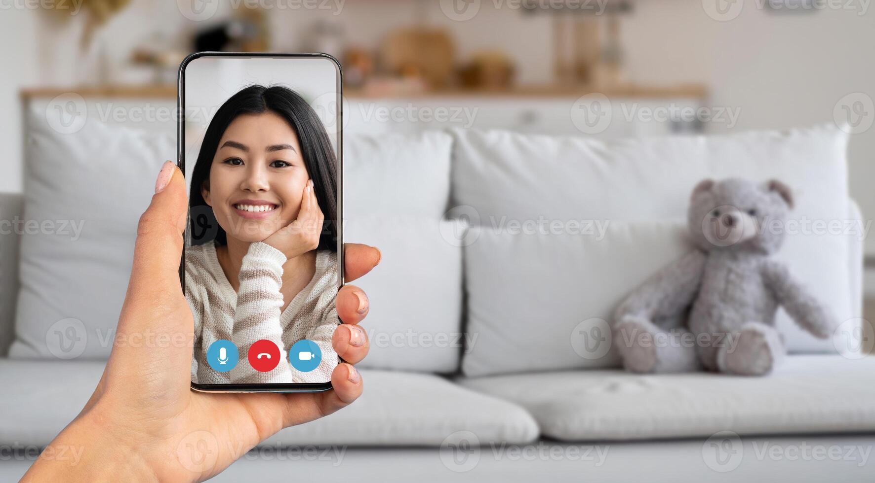 A person participating in a call from their home. They hold a smartphone showing a friendly face, surrounded by a cozy living room environment with soft furnishings. photo