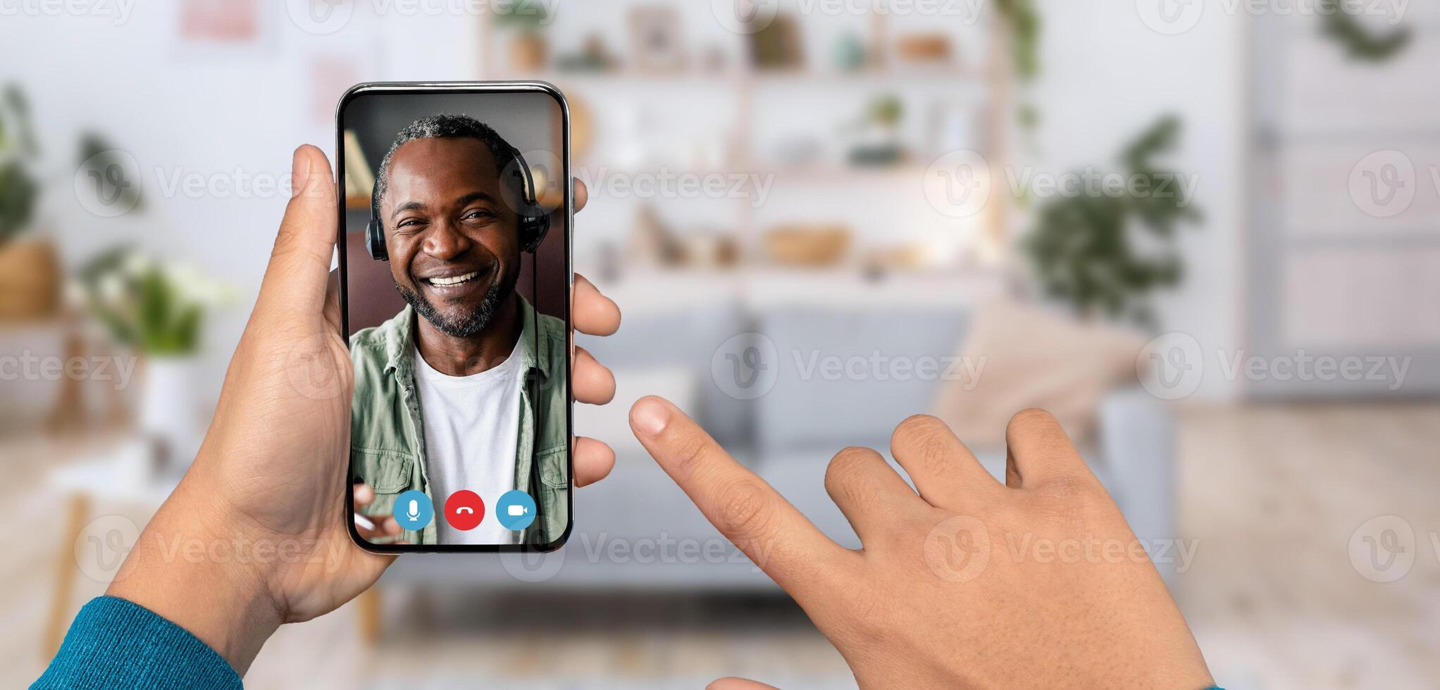 A person is using a smartphone for a call, smiling African America man is on the other end. The background shows a cozy home setting with plants and a couch. photo