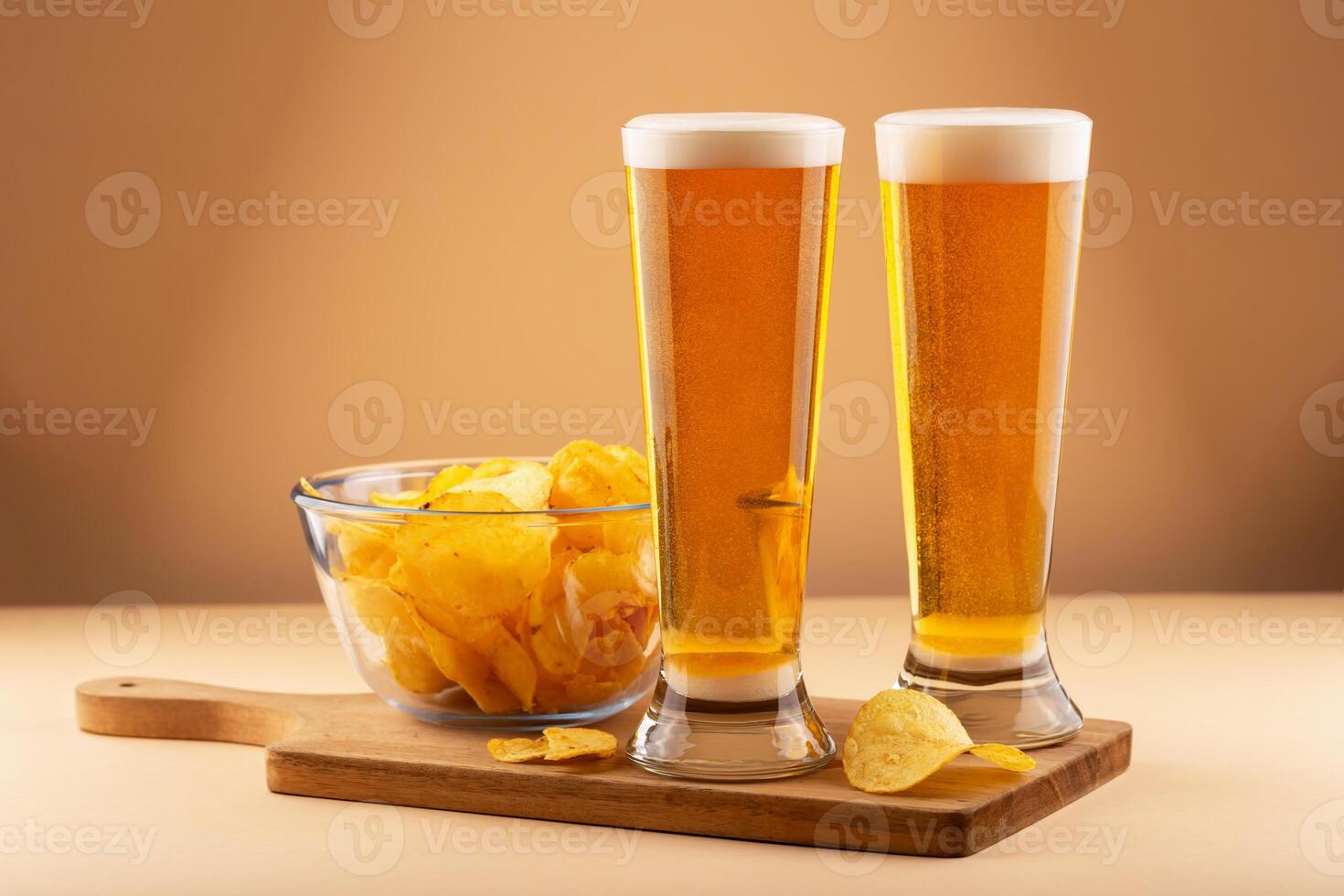 Two cold beer glasses and clear bowl of potato chips arranged on wooden board with warm background photo