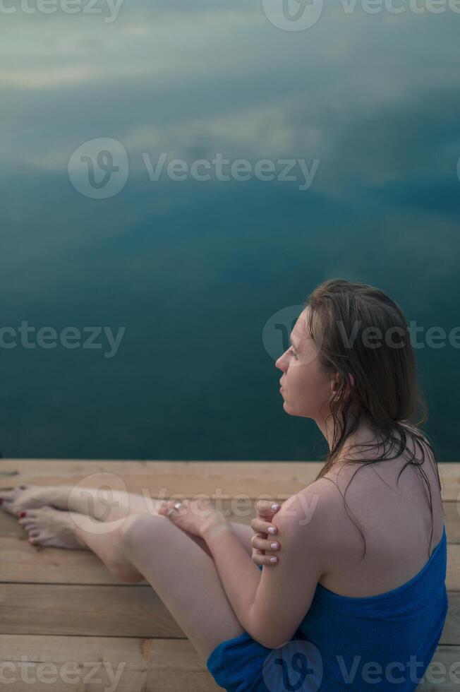 A young woman with wet hair, wrapped in a blue towel, sits on a wooden dock by a calm lake during sunset. photo