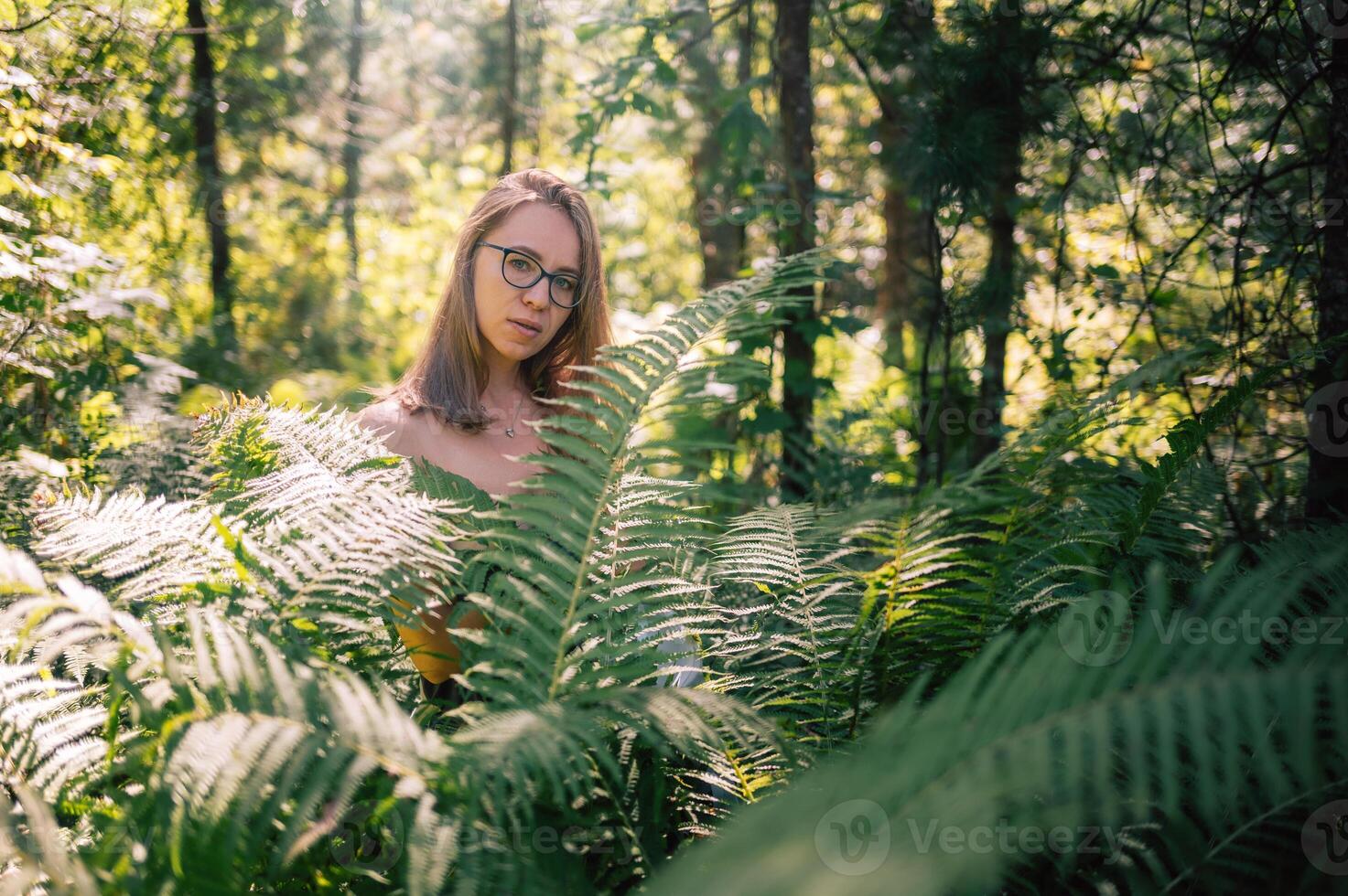 A young woman with glasses stands among vibrant green ferns in a sunlit forest, surrounded by nature and soft natural light. photo