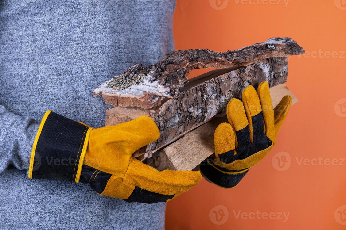 A person wearing yellow work gloves holds pieces of firewood against an orange background, emphasizing safety and outdoor work. photo