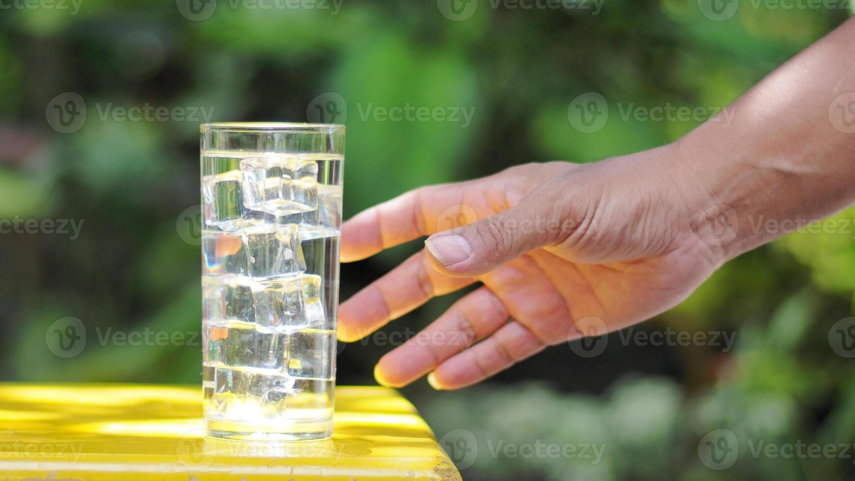 Hand Reaching for a Refreshing Glass of Ice Water on a Sunny Day with Greenery in the Background photo