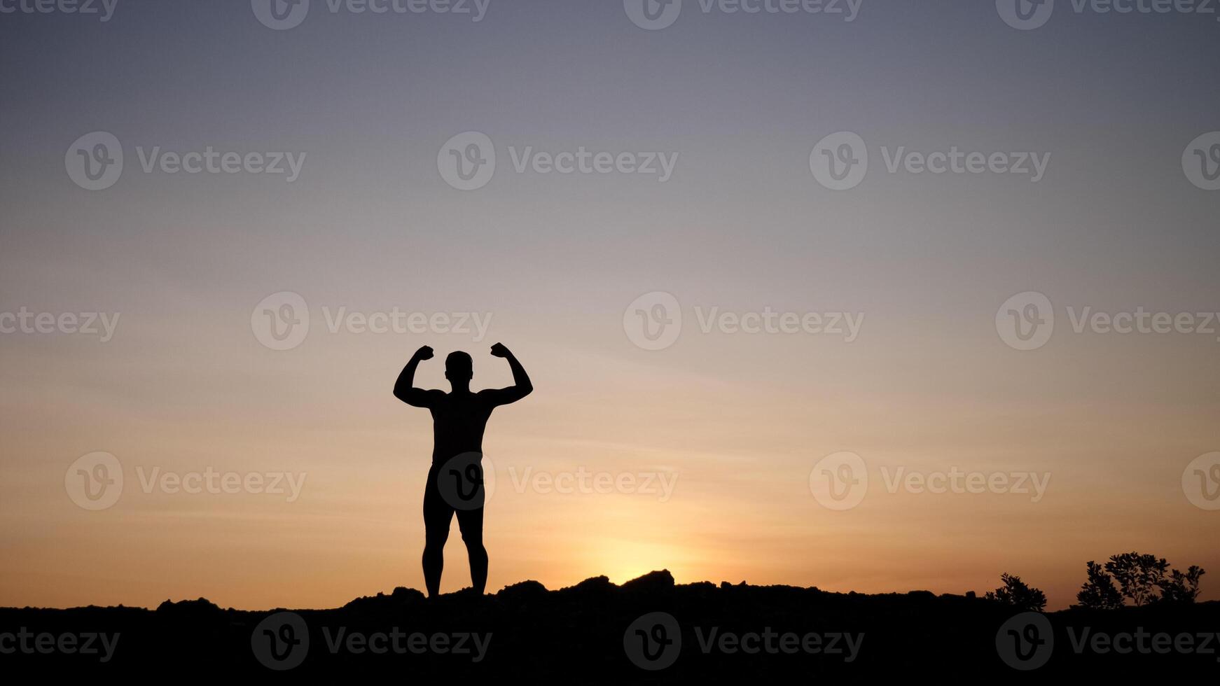 Silhouette of a Strong Man Flexing Muscles Against a Stunning Sunset Sky on a Rocky Hilltop photo