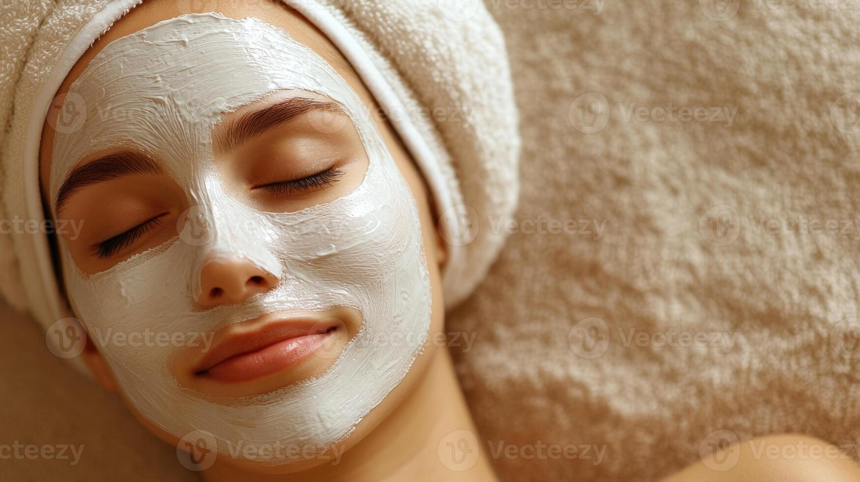 Young woman relaxing with white cosmetic mask on face during spa procedure. Eyes closed, towel wrapped head. Wellness and skincare treatment moment photo