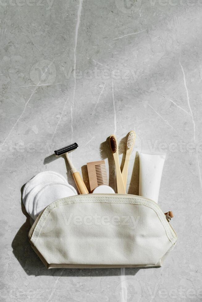 Hygiene products as bamboo toothbrushes, toothpaste, reusable pads, safety razor in toiletry bag on marble with natural light, eco-friendly items for self care, zero waste concept. Minimal style photo