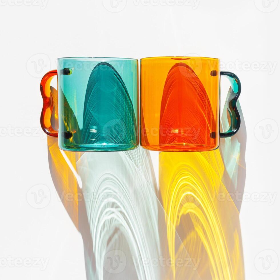 Two Modern Glass Cups with Vibrant sunlight Shadows, Aesthetic Top view of transparent orange and teal color glass mugs for drink. Yellow green reflections, shadow on white, minimal lifestyle photo