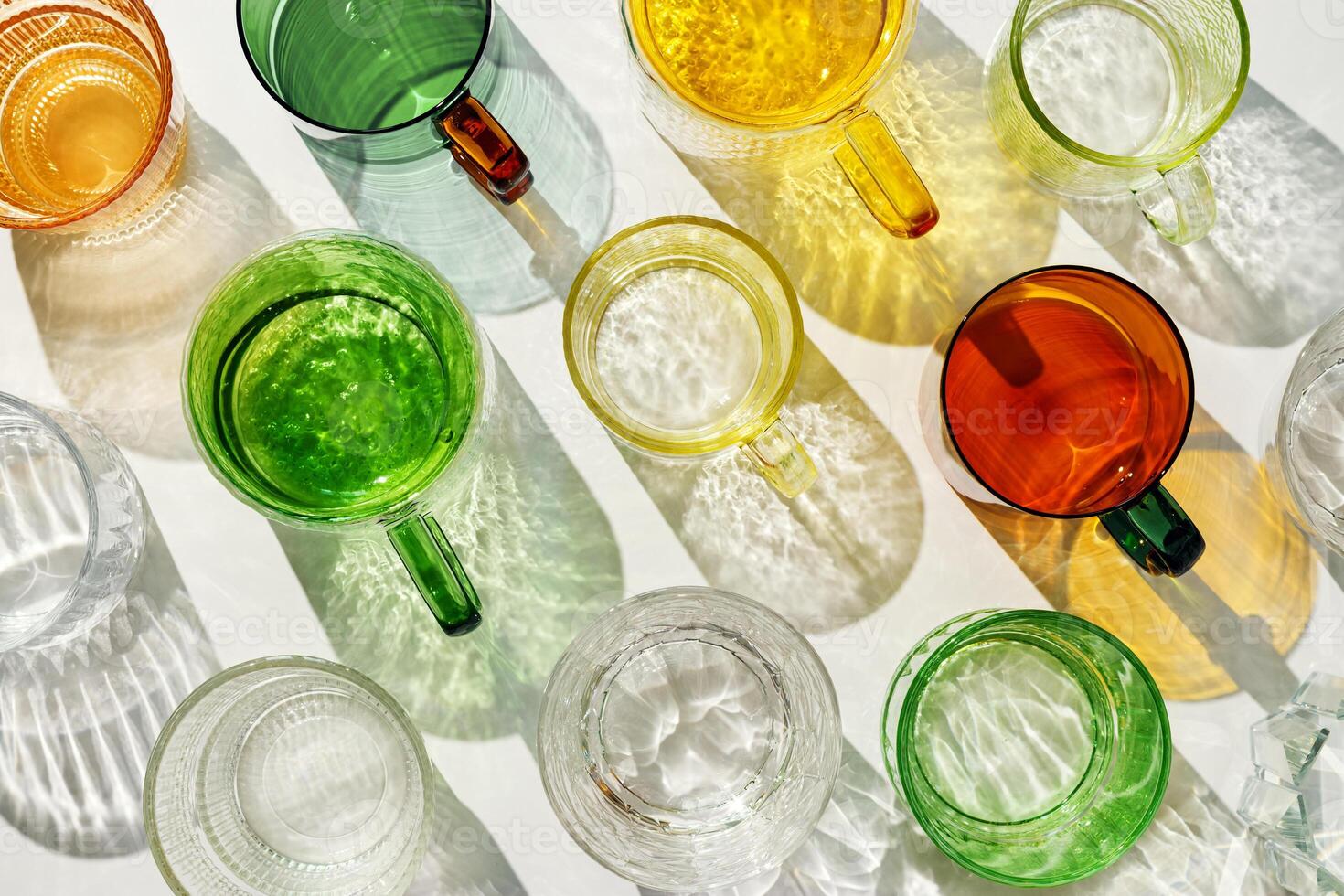 Vibrant set of colorful glass cups on white background, casting artistic light reflections and shadows in natural sunlight. Summer drink concept, minimal pattern of transparent glasses photo