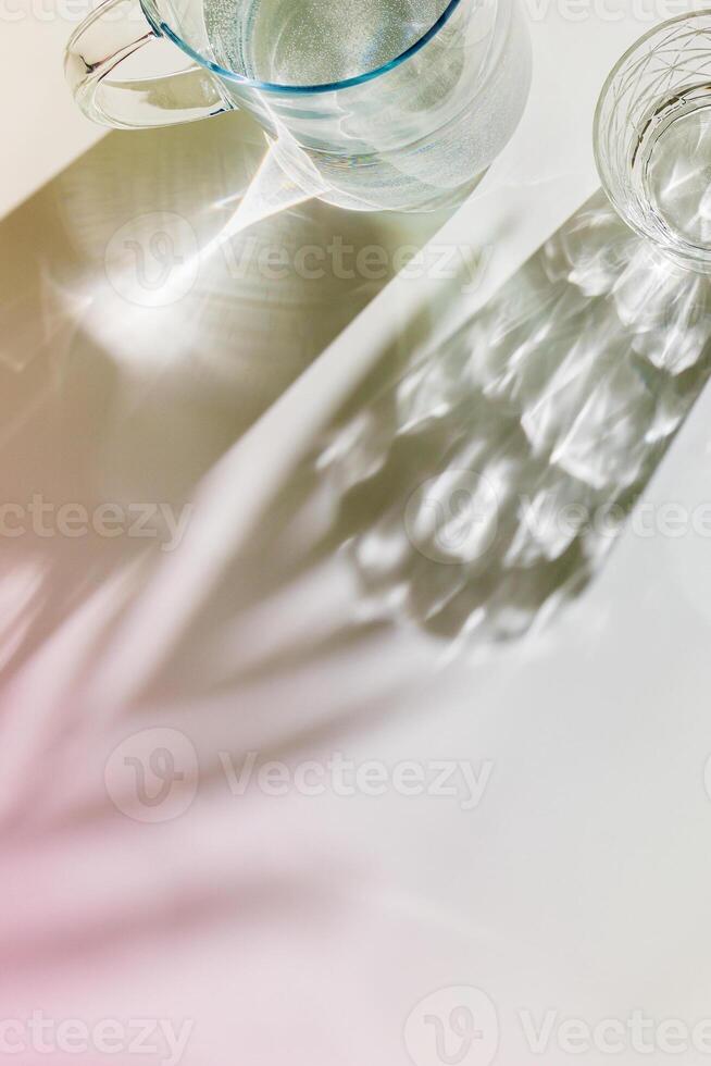 Transparent Water Glasses and water jug with palm leaf shadows at sunlight on white, pastel color photo minimal style, aesthetic top view shiny cups sparkling at natural light, summer drinks
