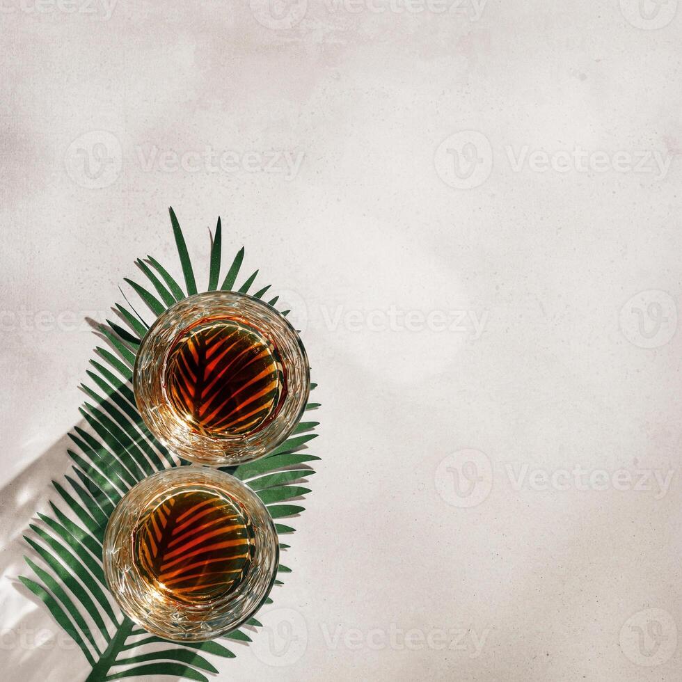 Whiskey Glasses with Shadows and sunlight Reflection on beige background, glass strong alcohol, palm leaf decor, aesthetic top view summer still life, refraction at crystal shiny cups, caustics photo