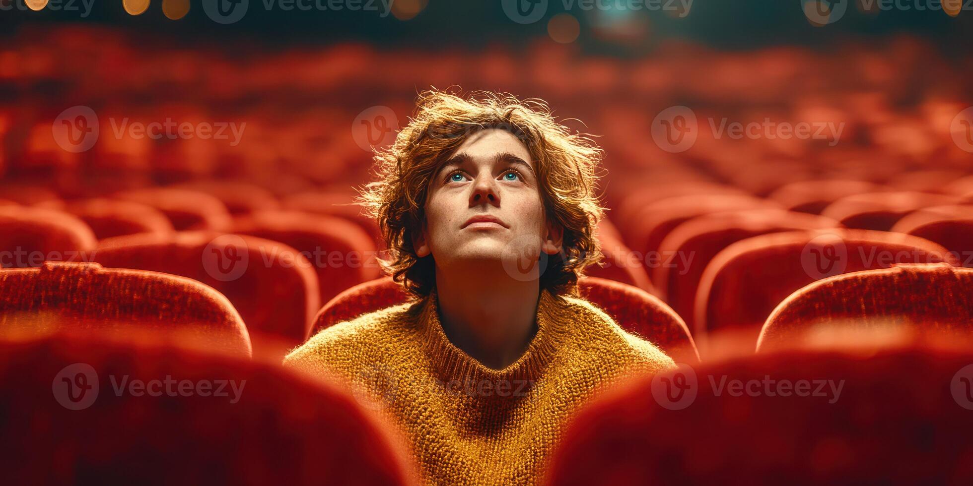 Young man gazes upward in a theater with red seats, lost in thought during a performance at dusk photo