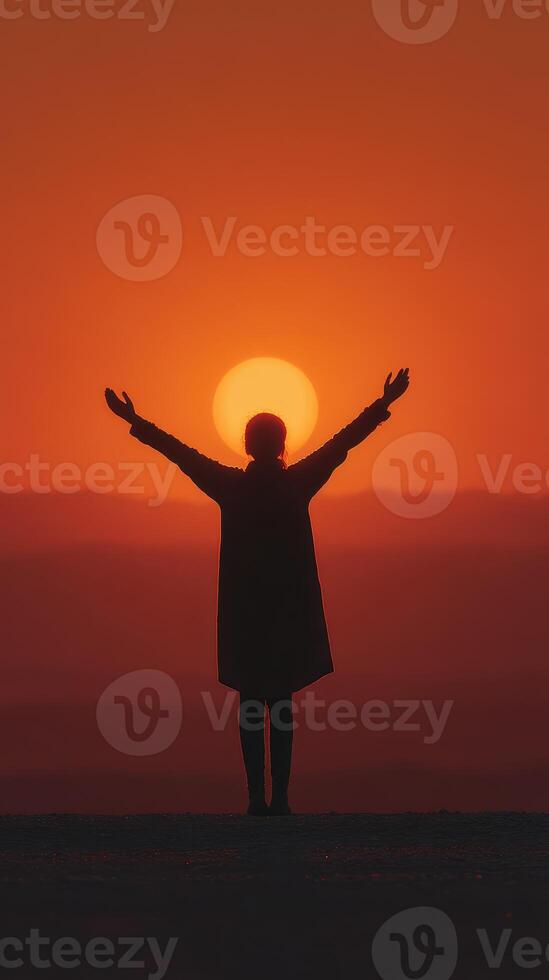 Person stands with arms raised in front of a sunset over distant hills during golden hour photo
