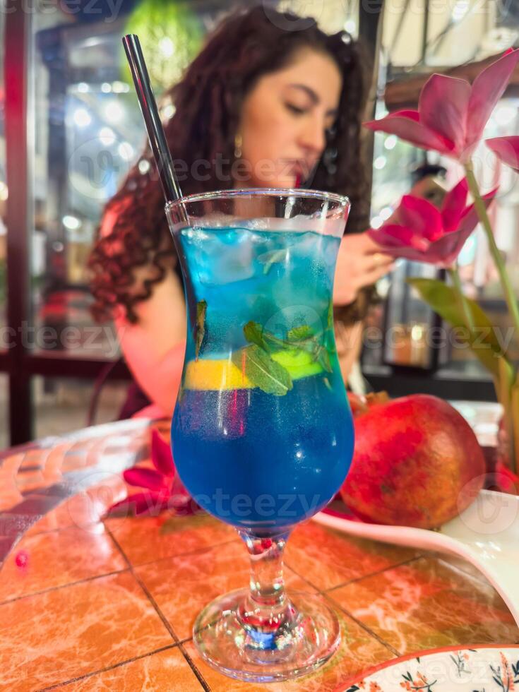 Blue bright mojito with black plastic tube in cafe on the street, against the background of a beautiful white girl with curly hair, vertical photo for social networks