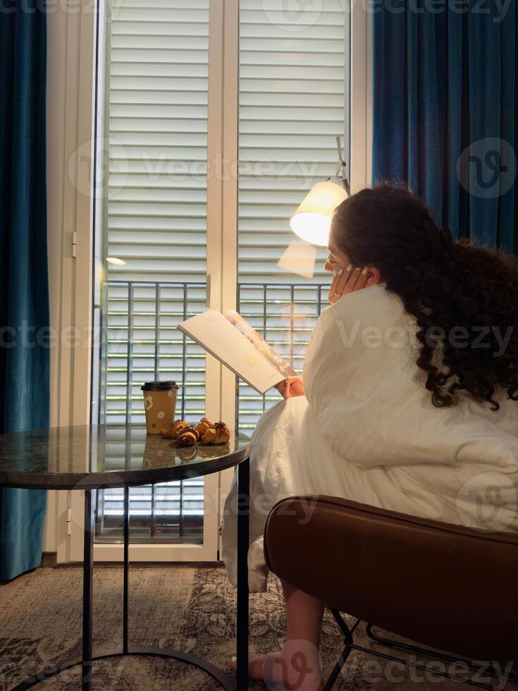 White girl reading magazine sitting in leather brown chair wrapped in white blanket near coffee table with coffee and croissants, vertical photo
