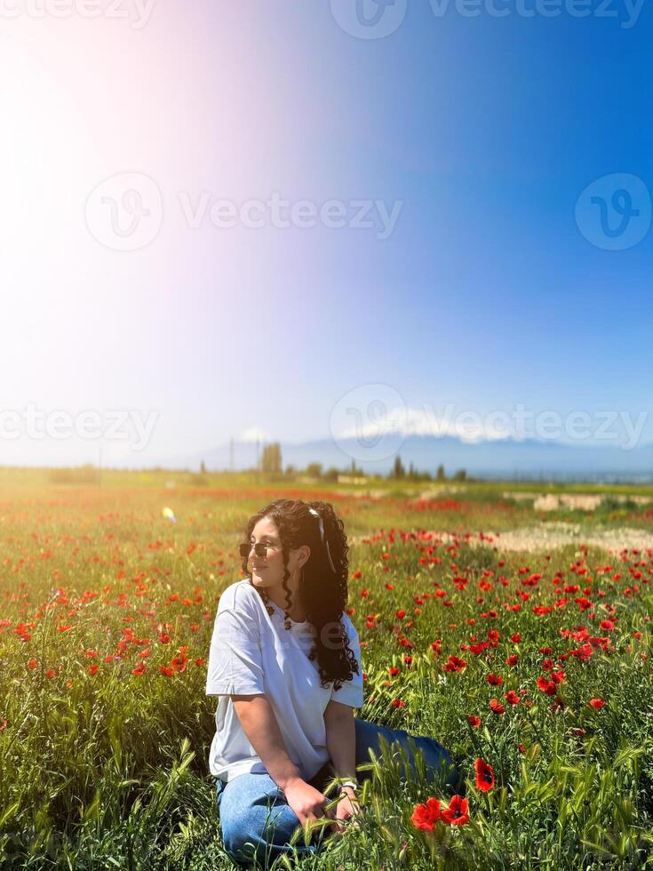 A white girl with curly hair is sitting in a field of red flowers against the background of Mount Ararat in Armenia on a bright sunny day, for an advertisement, for a banner, copy space photo